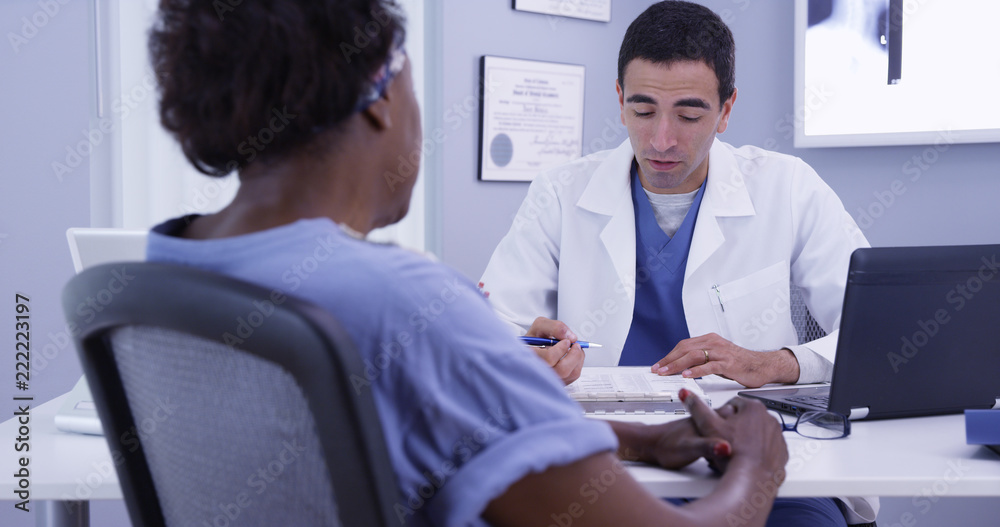 Happy senior patient talking with doctor in medical office about good health. Positive African woman meeting with young latino doctor to discuss her current health condition Happy senior patient talking with doctor in medical office about good health. Positive African woman meeting with young latino doctor to discuss her current health condition