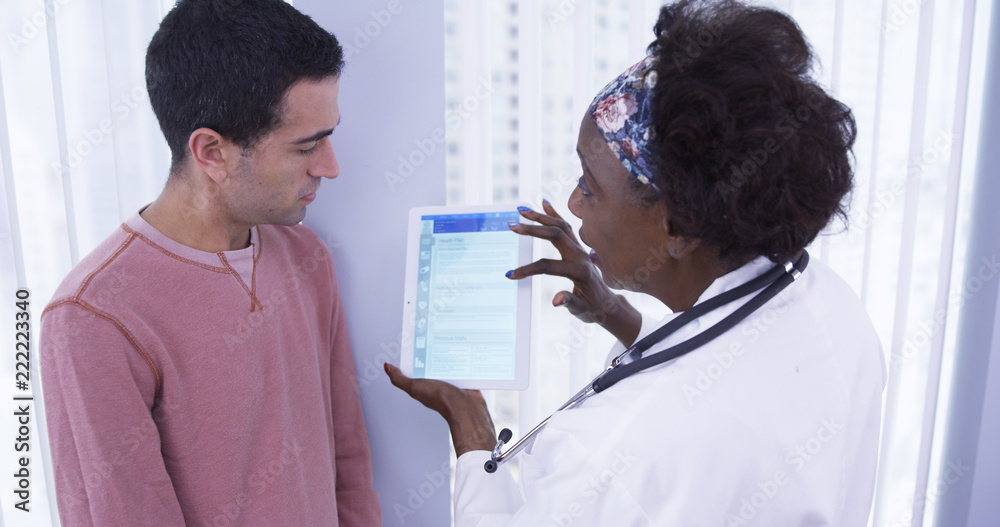 Obraz premium Mid aged black doctor using electronic notebook tablet to share different health plans with young male patient. Senior African MD showing male latino patient different health insurance plans on tablet