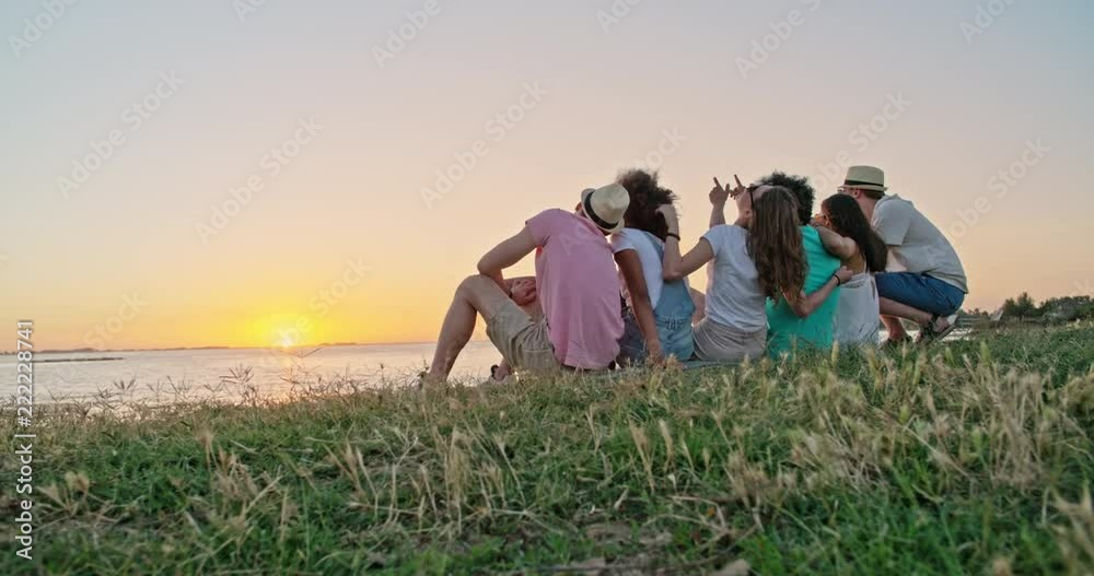 Six teen friends sitting on a meadow shore of a lake , meeting sunset and cheering, having fun 4k