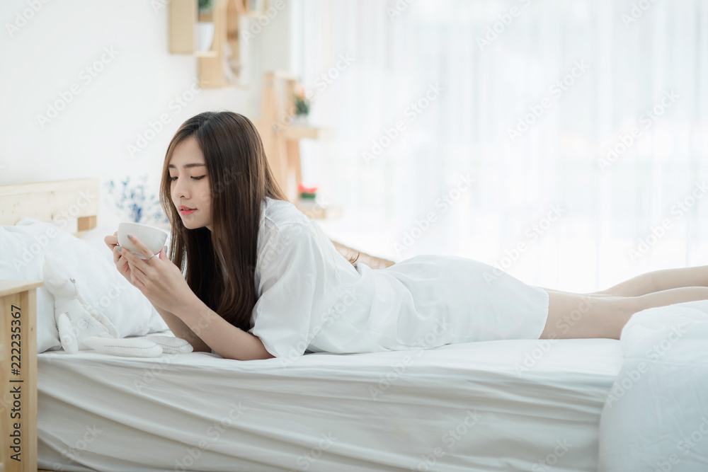 A beautiful young woman holding cup of coffee and lying on bed comfortably and happily. Happy morning. Portrait of a smiling pretty girl relaxing in white bed. A beautiful young woman holding cup of coffee and lying on bed comfortably and happily. Happy morning. Portrait of a smiling pretty girl relaxing in white bed.