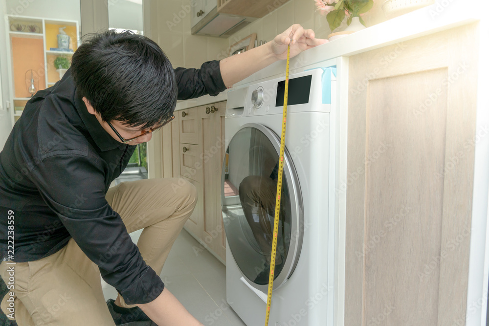 Young Asian worker man using tape measure for measuring washing machine in kitchen counter. Home interior design concept Young Asian worker man using tape measure for measuring washing machine in kitchen counter. Home interior design concept