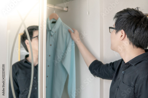 Young Asian man choosing casual style shirt in closet for dressing up in bedroom. Home living lifestyle concept