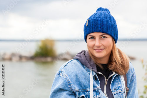 Portrait of outdoor atmospheric lifestyle photo of young beautiful  darkhaired woman  in knitting hat, in a denim jacket and black trousers against the background sea  in sunny autumn day