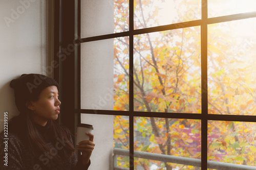 Asian beautiful woman thinking while drink coffee and standing in room with autumn leaf outside window background.Concept of foreign students homesick.