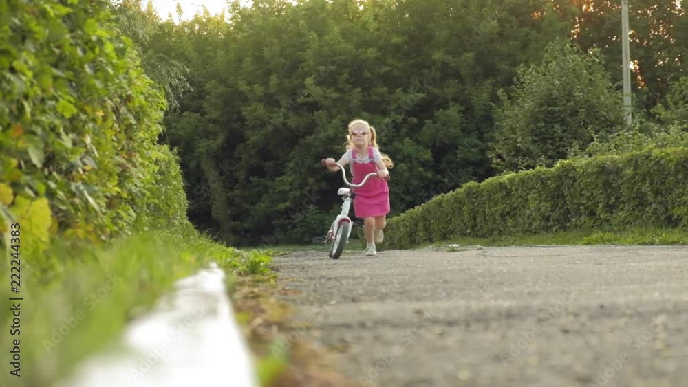 A happy, beautiful, little girl with long blond hair in a pink skirt and jumper rides a children's bike on the road, she smiles. Super slow motion A happy, beautiful, little girl with long blond hair in a pink skirt and jumper rides a children's bike on the road, she smiles. Super slow motion