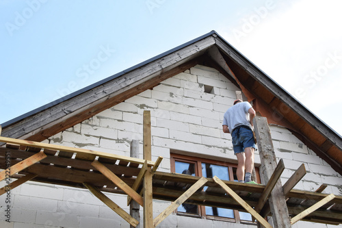Man installing wooden scaffolding next to unfinished lightweight concrete block house under construction in summer day. Home improvement concept.