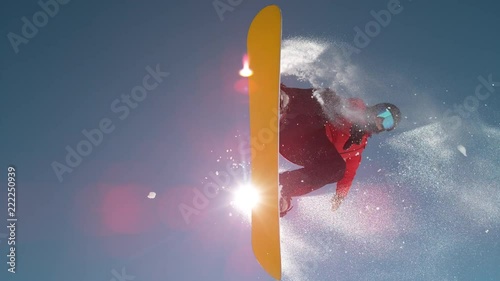 SLOW MOTION CLOSE UP: Snowboarder jumping big air kicker, spraying snowflakes and flying over sun on perfect winter day. Snowboard jump in snow park. Sunbeams shining past jumping boarder in mountains photo