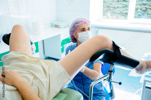 Female gynecologist in mask and hair cap with unrecognizable patient in gynecology chair looks on monitor while performing hysteroscopy operation. photo