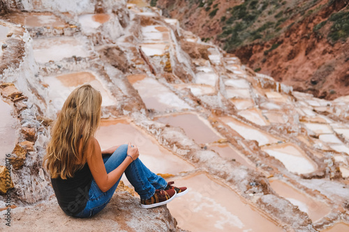 A blonde girl with flowing hair sits on the ground in jeans and looks at the salt water basins. Peru. Maras