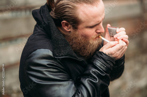 A bearded man in a black leather jacket stands outside and poses for a photographer. Smokes sigaret photo