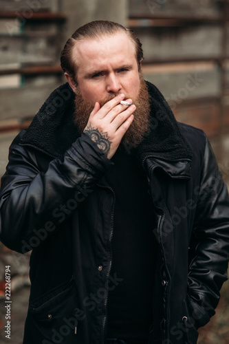A bearded man in a black leather jacket stands outside and poses for a photographer. Smokes sigaret