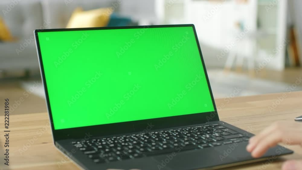 Man Uses Laptop with Green Mock-up Screen While Sitting at the Desk in His Cozy Living Room.