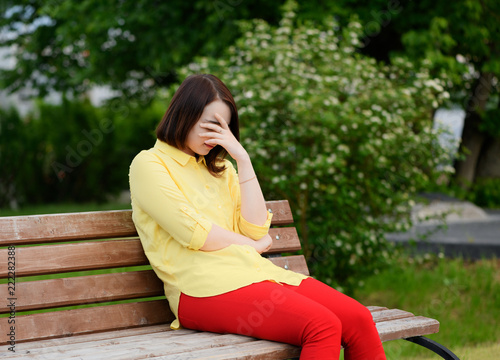 Valokuva The upset sad caucasian girl, 20 year old, is wearing the yellow shirt and the red pants sitting on a bench in a park