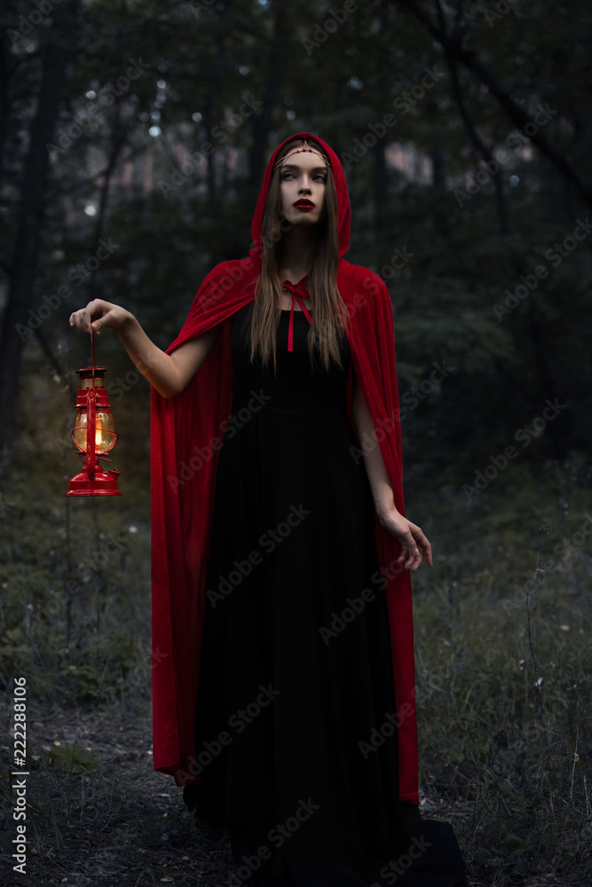 young mystic woman in red cloak with kerosene lamp walking in dark woods