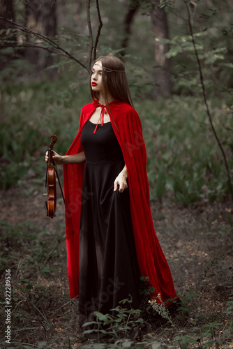 attractive mystic woman in red cloak holding violin in dark forest