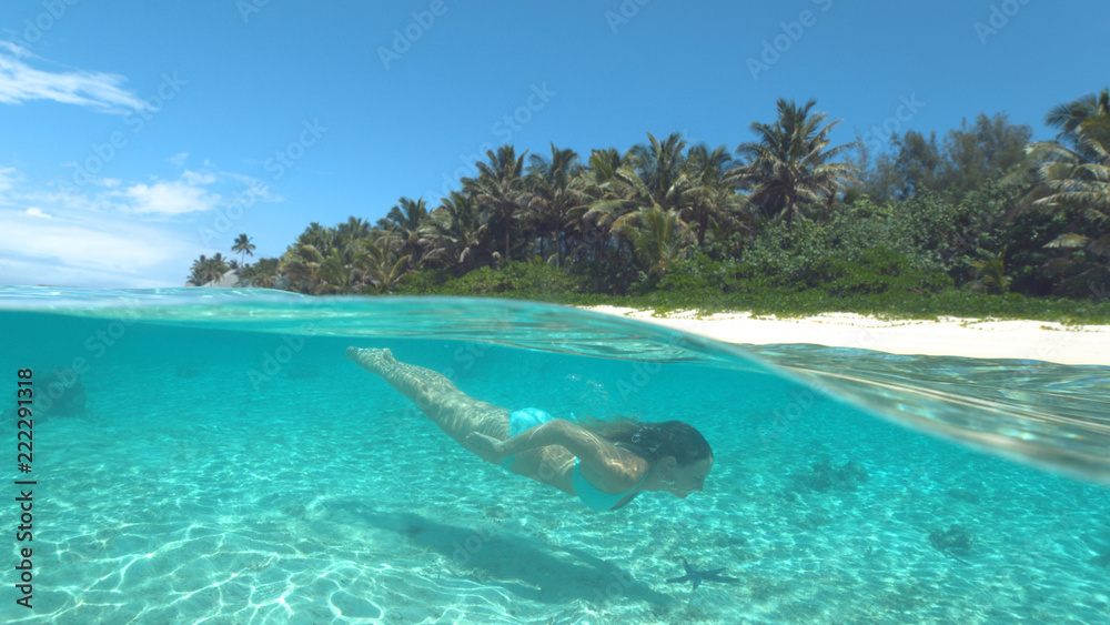 Fototapeta premium HALF UNDERWATER: Happy young woman in bikini diving near sunny tropical island.