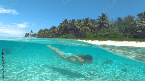 HALF UNDERWATER: Happy young woman in bikini diving near sunny tropical island.