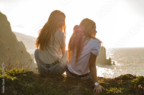 Two blond girls sitting on a cliff of island observing the horizon in a sunset of summer
