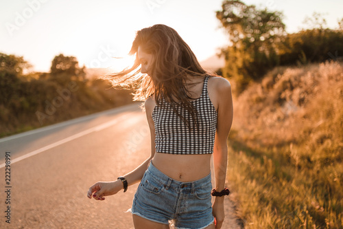 Young girl with hair bun and denim shorts dancing walking and smiling on the edge of the roadway in a sunset of summer