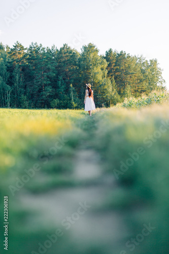 Fototapeta Naklejka Na Ścianę i Meble -  Girl walking on the field, in a hat and summer dress. Smiling and laughing, beautiful sunset in the forest and in nature. Happy traveler, lifestyle.