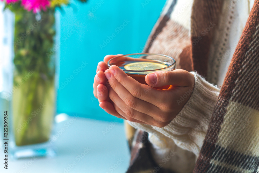 Obraz premium Tea drinking. The woman in a knitted, white pullover and in a warm plaid holds a cup of hot tea with lemon in her hands. Cozy weekends, winter drinks. Ceylon, Chinese tea