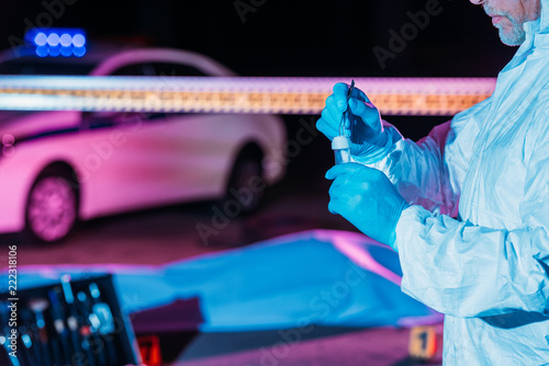 partial view of male criminologist in protective suit and latex gloves putting evidence into flask by tweezers at crime scene with corpse photo