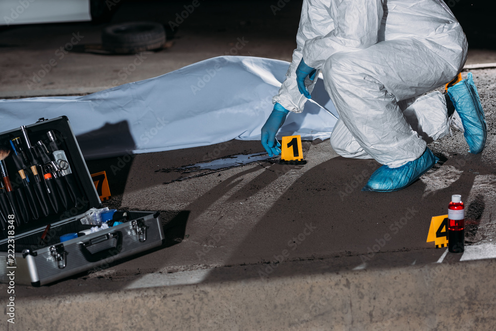 cropped image of criminologist in protective suit and latex gloves collecting evidence at crime scene with corpse