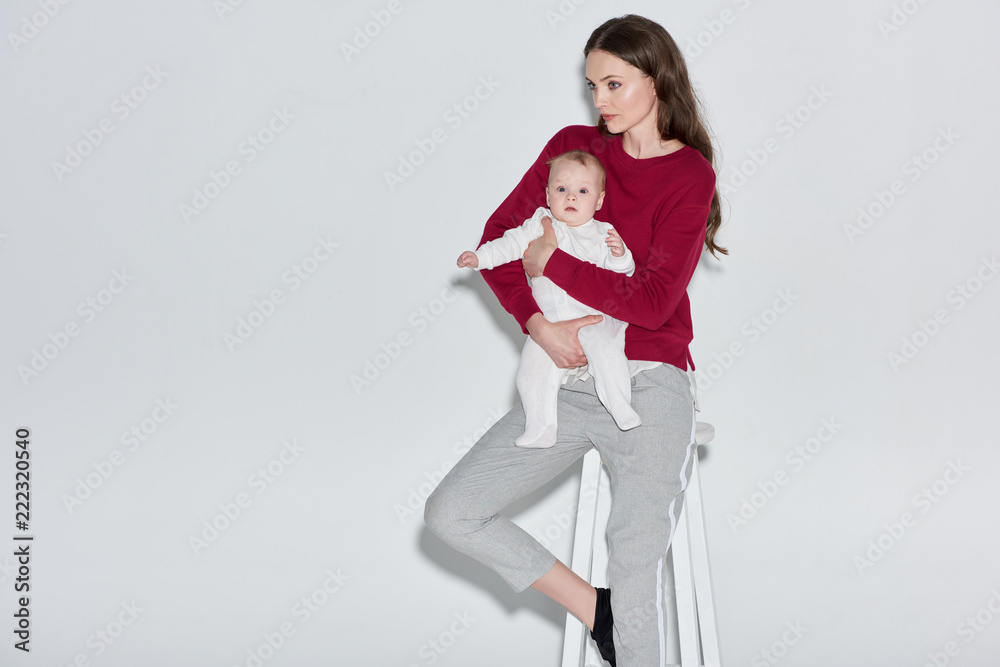 stylish young mother holding infant daughter and sitting on stool on grey stylish young mother holding infant daughter and sitting on stool on grey