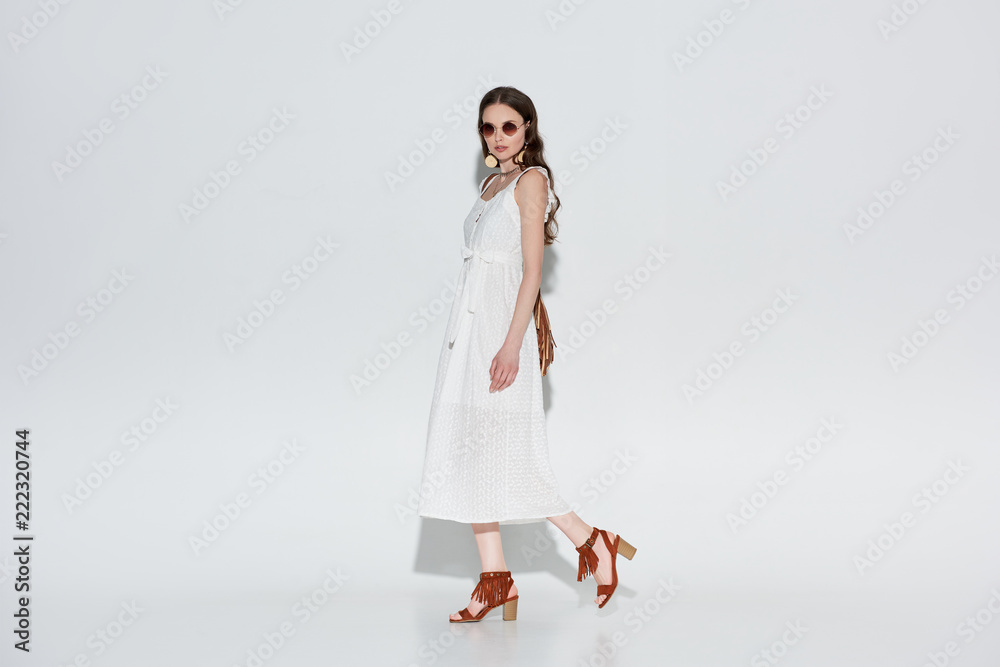 woman in stylish white dress and sunglasses holding handbag and walking on grey woman in stylish white dress and sunglasses holding handbag and walking on grey