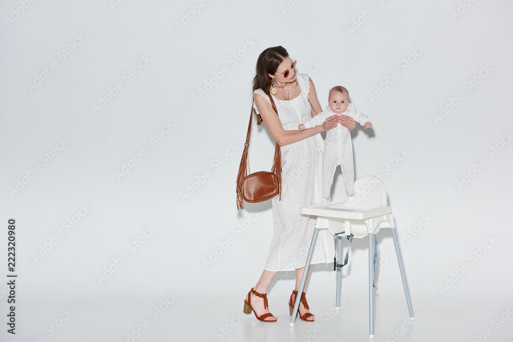 stylish woman in white dress and sunglasses carrying baby girl near high chair on grey