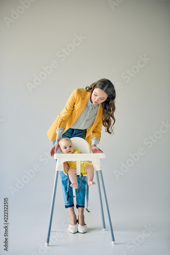fashionable mother looking at infant sitting in high chair on grey