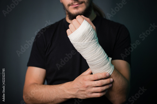 A young handsome guy in a black T-shirt with a bandaged hand on a gray background. A man with a hand in a bandage bandage.