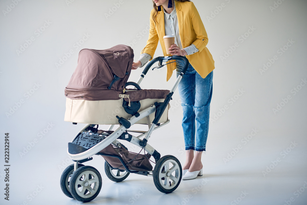 cropped shot of fashionable woman holding disposable coffee cup and standing with baby carriage on grey - obrazy, fototapety, plakaty 