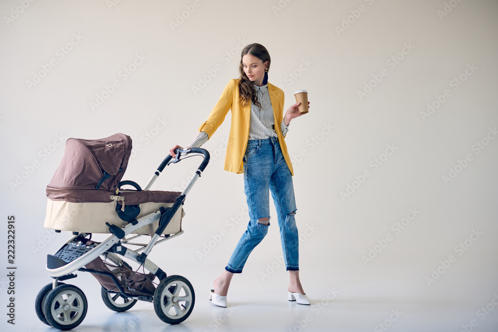 stylish young woman holding disposable coffee cup and looking at baby stroller on grey - obrazy, fototapety, plakaty 