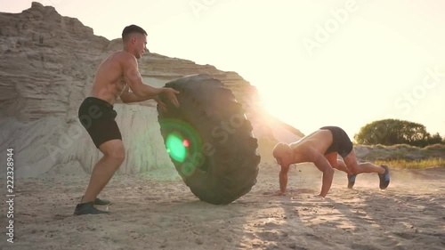 Two muscular open-chested athletes train in active mode on the beach doing push-UPS and pushing a huge wheel against a sandy mountain at sunset