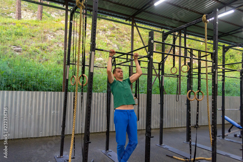 A young man in blue sports pants and a green T-shirt performs exercises pulling up on the horizontal bar in the gym outdoors with green grass on the background, rocky mountain, iron fence