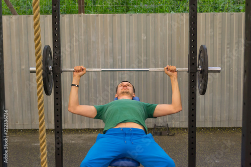 A man in a green T-shirt and blue pants doing a bar bench press, holding the dumbbell on his elbows, lying in the gym in the open air on the background of a rope, a metal fence and green bushes.