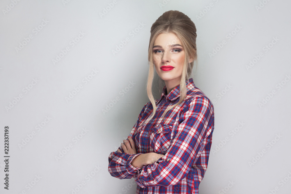 Satisfied beautiful blonde girl in pink checkered shirt, collected updo hairstyle and makeup standing, crossed hands and looking at camera and smiling. indoor studio shot, isolated on gray background Satisfied beautiful blonde girl in pink checkered shirt, collected updo hairstyle and makeup standing, crossed hands and looking at camera and smiling. indoor studio shot, isolated on gray background