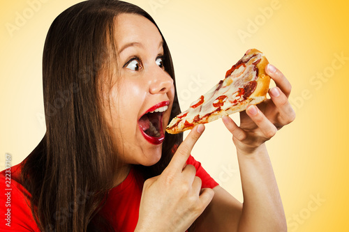 Young pretty girl eats piece of pizza with tomatoes  salami and cheese isolated on a yellow background.