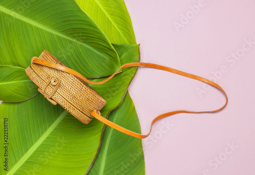 Fashionable handmade natural round rattan bag and tropical leaves on pink background flat lay. Top view with copy space. Trendy bamboo bag Ecobags from Bali. Summer fashion concept. photo