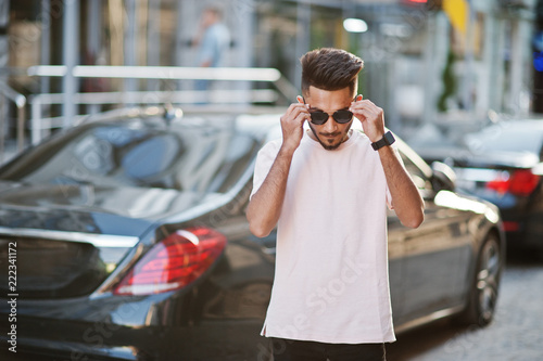 Stylish indian beard man at sunglasses and pink t-shirt against luxury car. India rich model posed outdoor at streets of city.