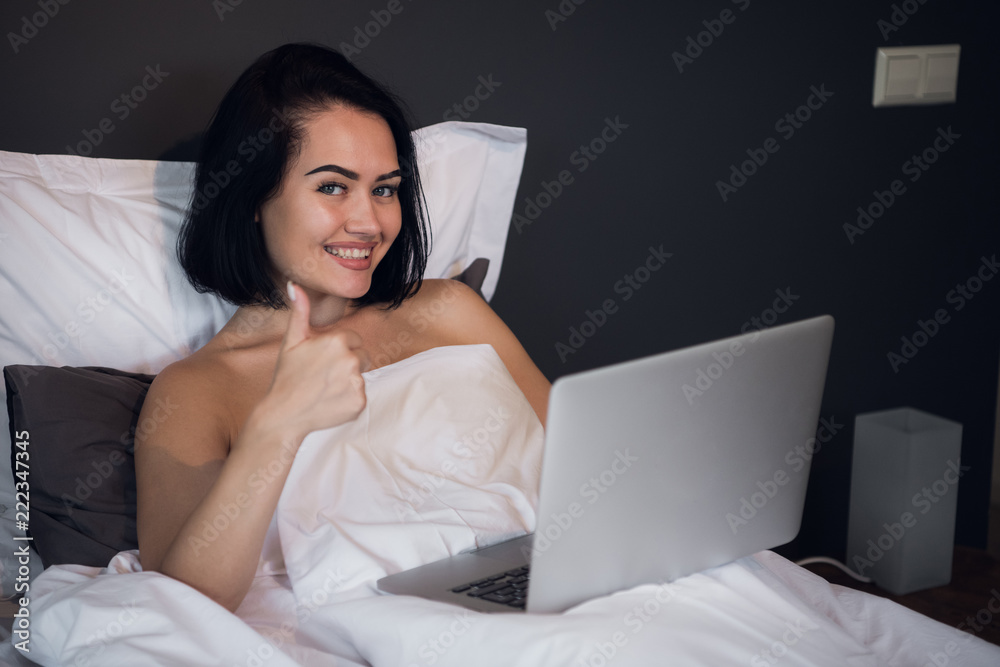 happy woman typing on her laptop and showing thumbs up while lying in a bed at home happy woman typing on her laptop and showing thumbs up while lying in a bed at home