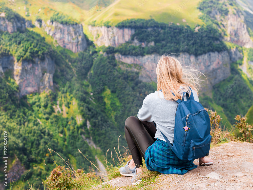 Naklejka premium A girl sits on the edge of the cliff and looking at the valley. Woman enjoying the forest view from the top of the mountain in summer. The traveler girl at the peak of mountain.