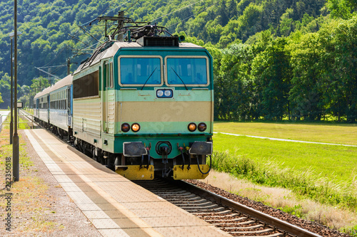 A green electric locomotive passing the Czech countryside. A train running through the green valley. Rail transport in the Czech Republic. The train arriving on a rural platform