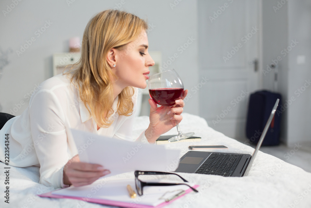 Cheerful business woman holding glass of wine in bed. Reading report and looking at laptop. Business in bed concept. Cheerful business woman holding glass of wine in bed. Reading report and looking at laptop. Business in bed concept.
