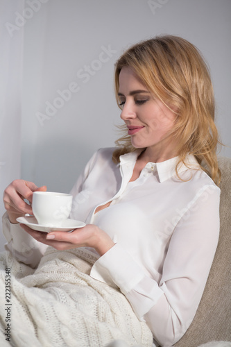 Blond businesswoman having coffee while sitting on white arm chair. Isolated on white background.