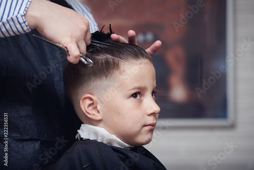 Barber making hairstyle to a Caucasian boy using scissors and hairbrush.