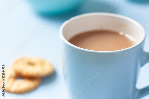 A blue mug of tea and biscuits with tea pot and accessories on a pale blue background