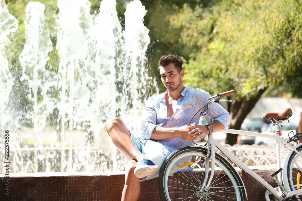 Obraz premium Handsome young hipster man sitting with bicycle near fountain in park