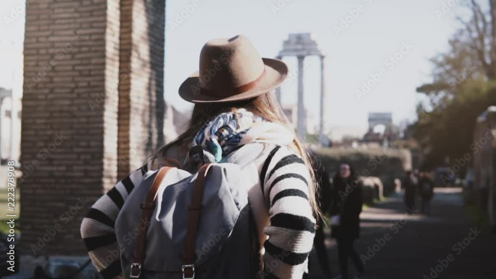 Camera moving around young tourist woman in hat with backpack and arms on hips standing at Forum of Rome ruins in Italy.